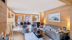 Un salon moderne avec un canapé, une table basse et des fauteuils. On y trouve également un coin repas avec une table et des chaises. Les murs de la pièce sont en bois et elle est dotée d’une grande fenêtre.
