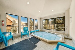 Un espace détente avec un jacuzzi, deux fauteuils bleus et de grandes fenêtres donnant sur des arbres et des montagnes enneigés.