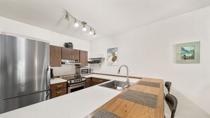 A modern kitchen with a stainless steel refrigerator, microwave, stove, and sink. There are wooden cabinets and a white countertop. The kitchen has a wooden island with seating and a sink. - Newly Renovated, One Bedroom Condominium at Purgatory Steps From Slopes (Durango)