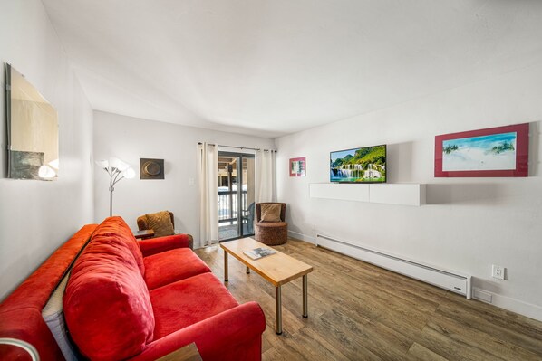 Un salon moderne avec un canapé rouge, une table basse en bois et une vue sur l’extérieur grâce à une porte vitrée.