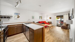 A modern kitchen with a central island, wooden cabinets, and a built-in dishwasher. There is a red sofa and a small table in the living area, with a view of the outdoors through a sliding glass door. - Newly Renovated, One Bedroom Condominium at Purgatory Steps From Slopes (Durango)