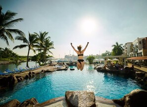 En person som hoppar i en pool omgiven av palmer och solstolar.