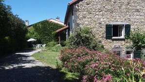 Une maison en pierre avec une allée gravillonnée, un mur en pierre et un jardin agrémenté de fleurs roses.