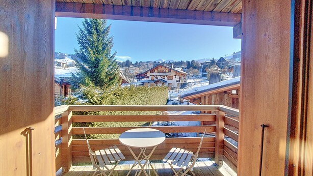 Un balcón de madera con una mesa y sillas, con vistas a un pueblo nevado con casas y un alto árbol de hoja perenne.