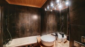 Un baño moderno con baldosas oscuras, una bañera blanca, un espejo grande y una zona de ducha con iluminación superior.