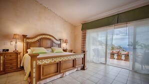Une chambre avec un grand lit, une tête de lit en bois, deux tables de chevet avec des lampes et une vue sur un balcon agrémenté de plantes en pot.