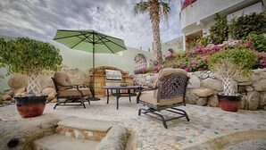 Une terrasse extérieure avec un sol en pierre, un barbecue et deux fauteuils en osier accompagnés d’une table et d’un parasol.