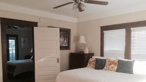 Ein Schlafzimmer mit Bett, Nachttisch, Deckenventilator, Fenster mit Jalousien und einer Tür zu einem anderen Raum.