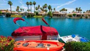 A poolside area with a covered kayak and a paddle boat, palm trees, and a building in the background. - Executive Lakefront La Quinta Gem with Infinity Pool, Boats, and Casita (La Quinta)