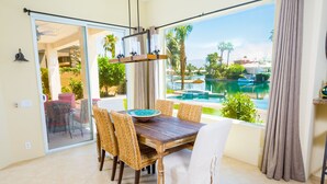 A dining area with a wooden table and wicker chairs, a large window with a view of a swimming pool and palm trees, and a sliding glass door leading to an outdoor patio. - Executive Lakefront La Quinta Gem with Infinity Pool, Boats, and Casita (La Quinta)