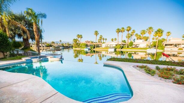 A swimming pool with a curved edge, surrounded by palm trees and a clear blue sky. - Executive Lakefront La Quinta Gem with Infinity Pool, Boats, and Casita (La Quinta)