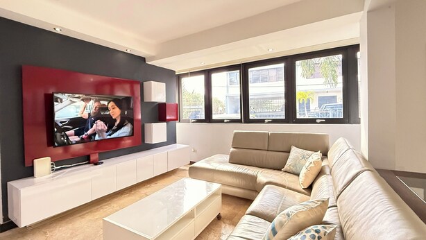 Een moderne woonkamer met een grote flatscreen-tv aan een rode muur, een beige bank met gekleurde kussens, een witte salontafel en een witte dressoir met opbergruimte.
