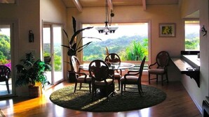 Un espace repas comprenant une table ronde et des chaises, un ventilateur de plafond et de grandes fenêtres donnant sur un paysage verdoyant.