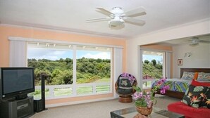 Un salon spacieux doté d’une grande fenêtre, d’un ventilateur de plafond, d’une télévision, d’un canapé et d’un lit recouvert d’une couvre-lit aux couleurs vives.