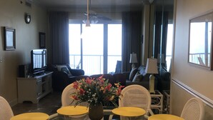 Ein Hotelzimmer mit Essbereich, ausgestattet mit Korbstühlen, einem Granittisch, einer Couch, einem Fernseher und einem großen Fenster mit Vorhängen.