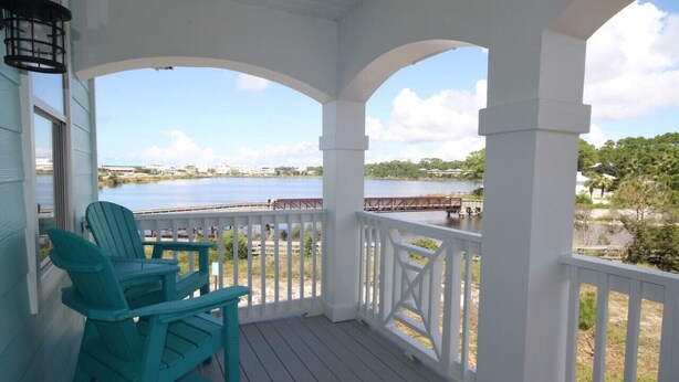 Eine überdachte Veranda mit weißen Geländern und türkisfarbenen Adirondack-Stühlen, mit Blick auf einen stillen See und eine entfernte Brücke.