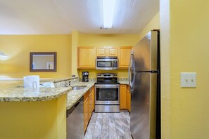 A compact kitchen with granite countertops, wooden cabinets, a stainless steel refrigerator, a microwave, an oven, and a coffee maker. - Harbour Lights™ - 1 Bedroom Deluxe (Myrtle Beach)