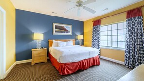 A hotel room with a bed, two bedside tables, a ceiling fan, a window with patterned curtains, and a painting on the wall. - Harbour Lights™ - 1 Bedroom Deluxe (Myrtle Beach)