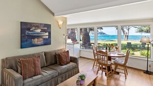 Una sala de estar con sofá, una mesa de centro y un comedor con sillas y mesa. Hay un cuadro en la pared y vistas al océano a través de amplias ventanas.