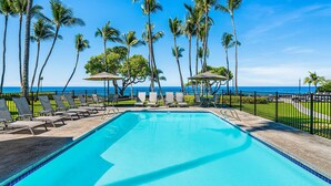 A pool area with lounge chairs and umbrellas, surrounded by palm trees and a view of the ocean. - 🏖️Desirable Ground Floor Unit | AC & WIFI | Concierge Included (Kailua Kona)