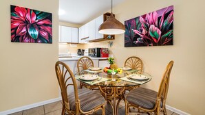 A dining area with a glass table surrounded by wicker chairs, a hanging pendant light, and two vibrant floral paintings on the walls. - 🏖️Desirable Ground Floor Unit | AC & WIFI | Concierge Included (Kailua Kona)