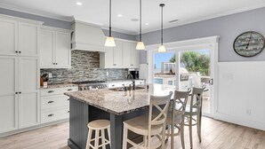 Una cocina moderna con armarios blancos, una isla grande con taburetes de bar, iluminación colgante y vistas a un patio exterior.