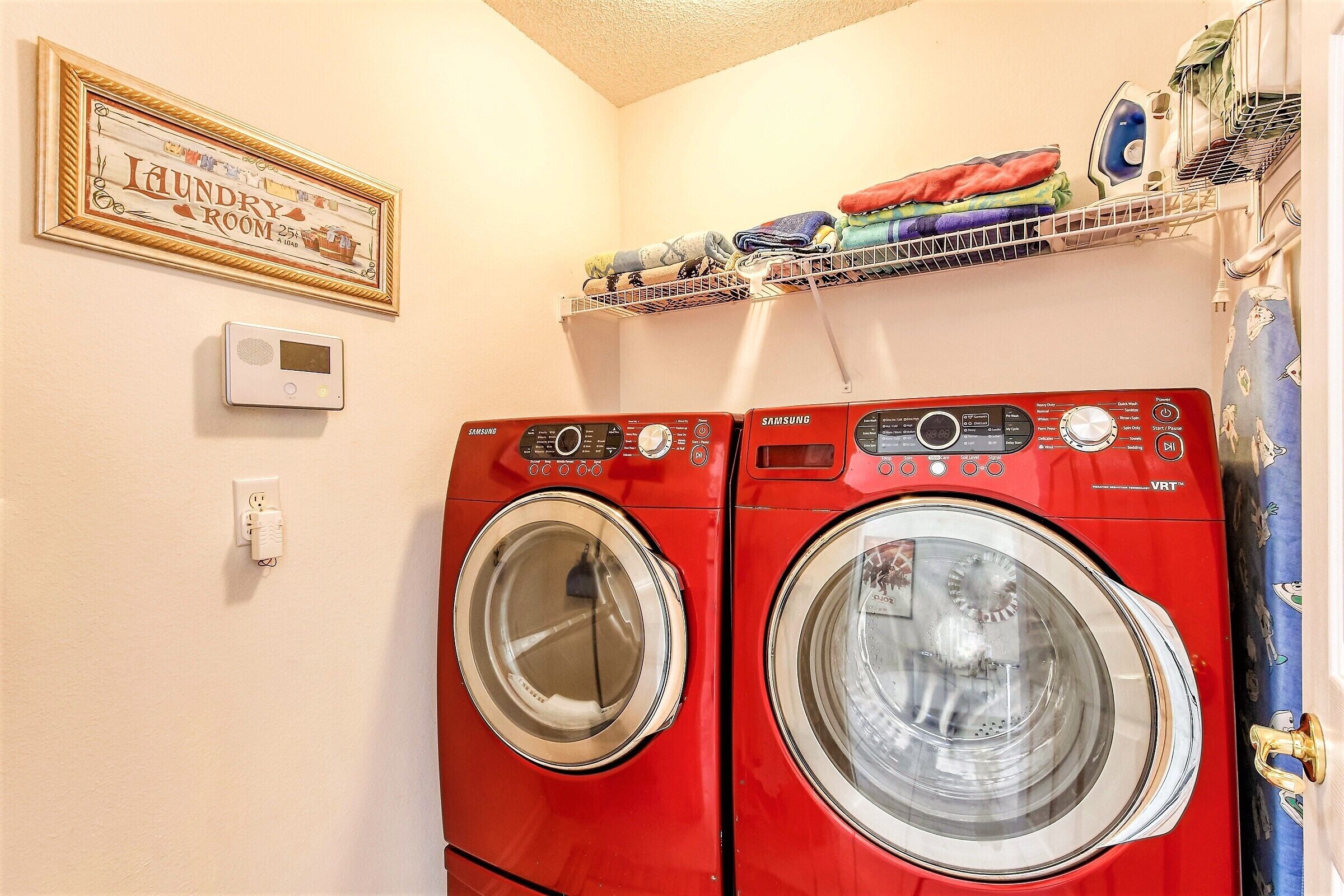 Une buanderie équipée de deux machines à laver à chargement frontal de couleur rouge, d’une étagère où le linge plié est rangé, d’un thermostat fixé au mur et d’une pancarte encadrée placée au-dessus des machines à laver.