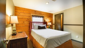 A hotel room with a wooden wall, a bed with white bedding, a nightstand with a lamp, and a door. - Wisconsin Dells | Pool & Waterpark Access | 2BR Timber (Wisonsin Dells)