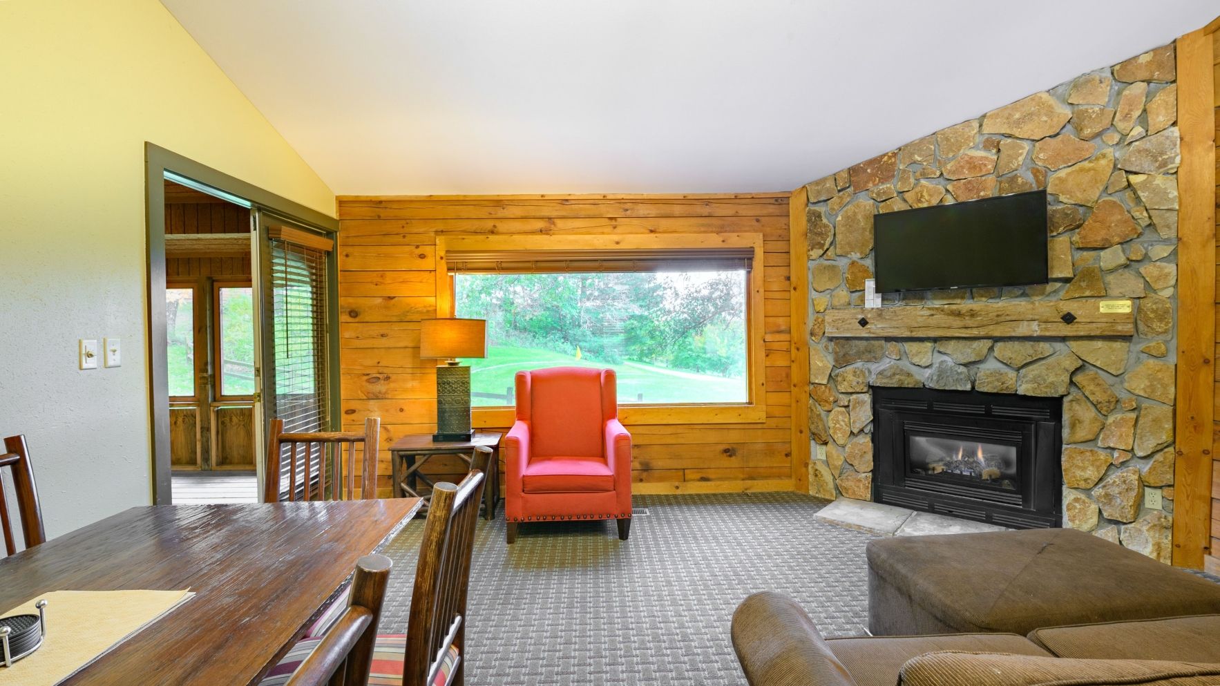 A cozy cabin interior with a stone fireplace, a red chair, a dining table, and a television mounted on the stone wall.