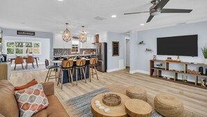 Una sala moderna con sofá, mesa de centro y mueble para televisión. Se ve una cocina con comedor al fondo.