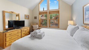 A bedroom with a large bed, a television on a wooden dresser, a chair, and a window with a view of a house. - Keystone Townhome Between River Run & Mountain House & Close to Shops/Dining (Keystone)