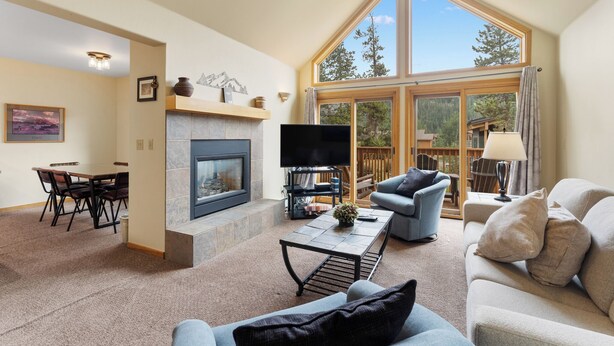 A spacious living room with a fireplace, a television, and a large window offering a view of the outdoors. - Keystone Townhome Between River Run & Mountain House & Close to Shops/Dining (Keystone)