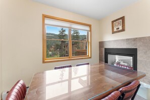 A dining area with a large glass table, leather chairs, a fireplace, and a window with a view of trees and a cabin. - Keystone Townhome Between River Run & Mountain House & Close to Shops/Dining (Keystone)