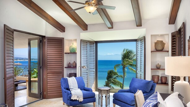 Ein modernes Wohnzimmer mit Blick aufs Meer, ausgestattet mit zwei blauen Sesseln, einem kleinen Beistelltisch und einem Deckenventilator.