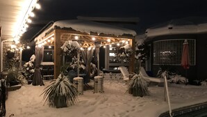 Área externa coberta de neve com um gazebo de madeira, luzes de cordão e cadeiras de descanso.