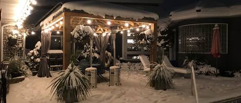 Un espacio exterior cubierto de nieve con un quiosco de madera, luces colgantes y sillones de descanso.