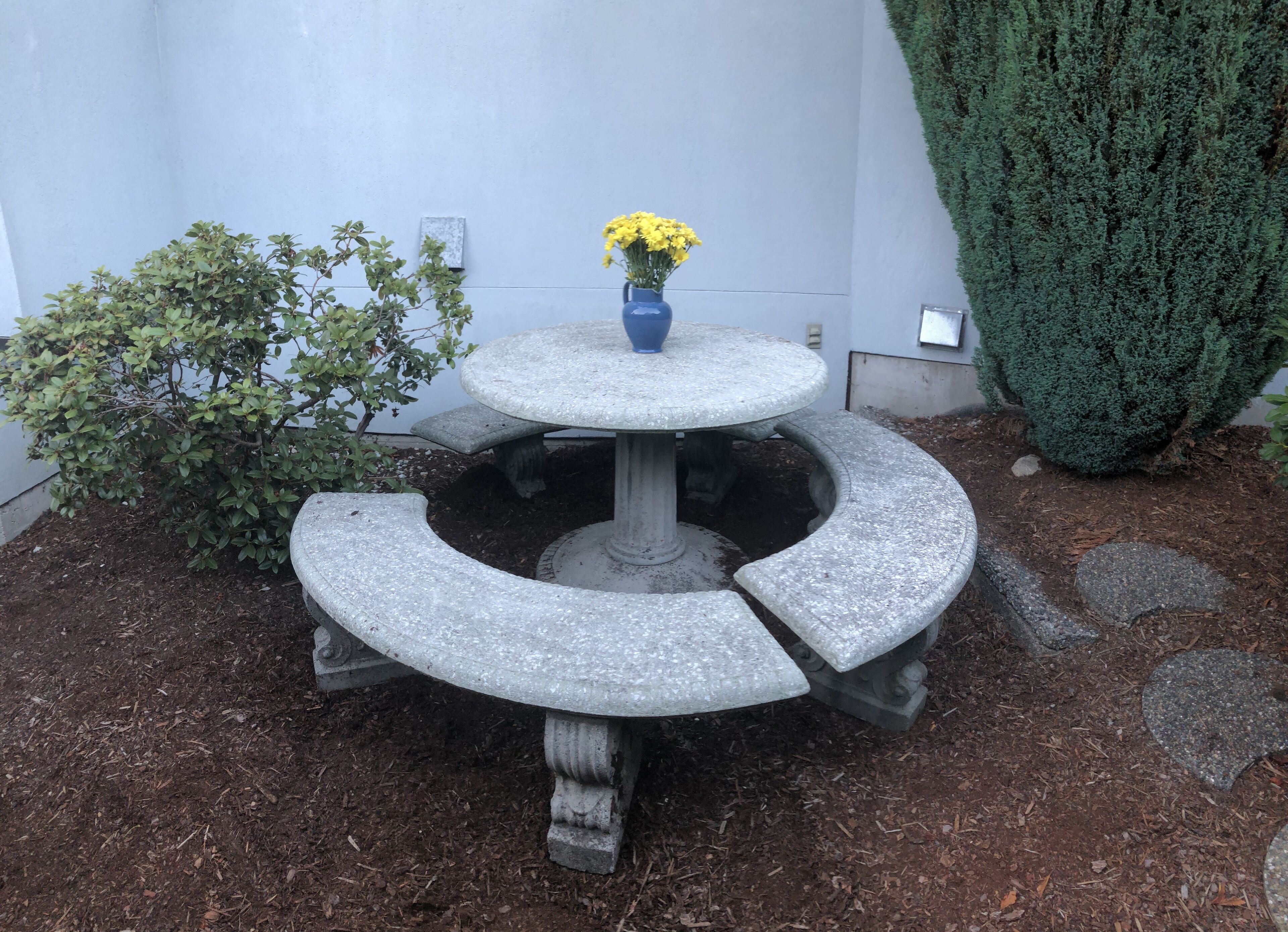 Une fontaine en pierre, ornée d’un vase rempli de fleurs jaunes, entourée de paillis et d’un petit arbuste.