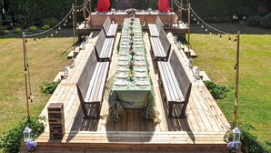 Uma área de refeições ao ar livre com uma mesa comprida preparada para vários convidados, cercada por bancos de madeira e guarda-sóis vermelhos.