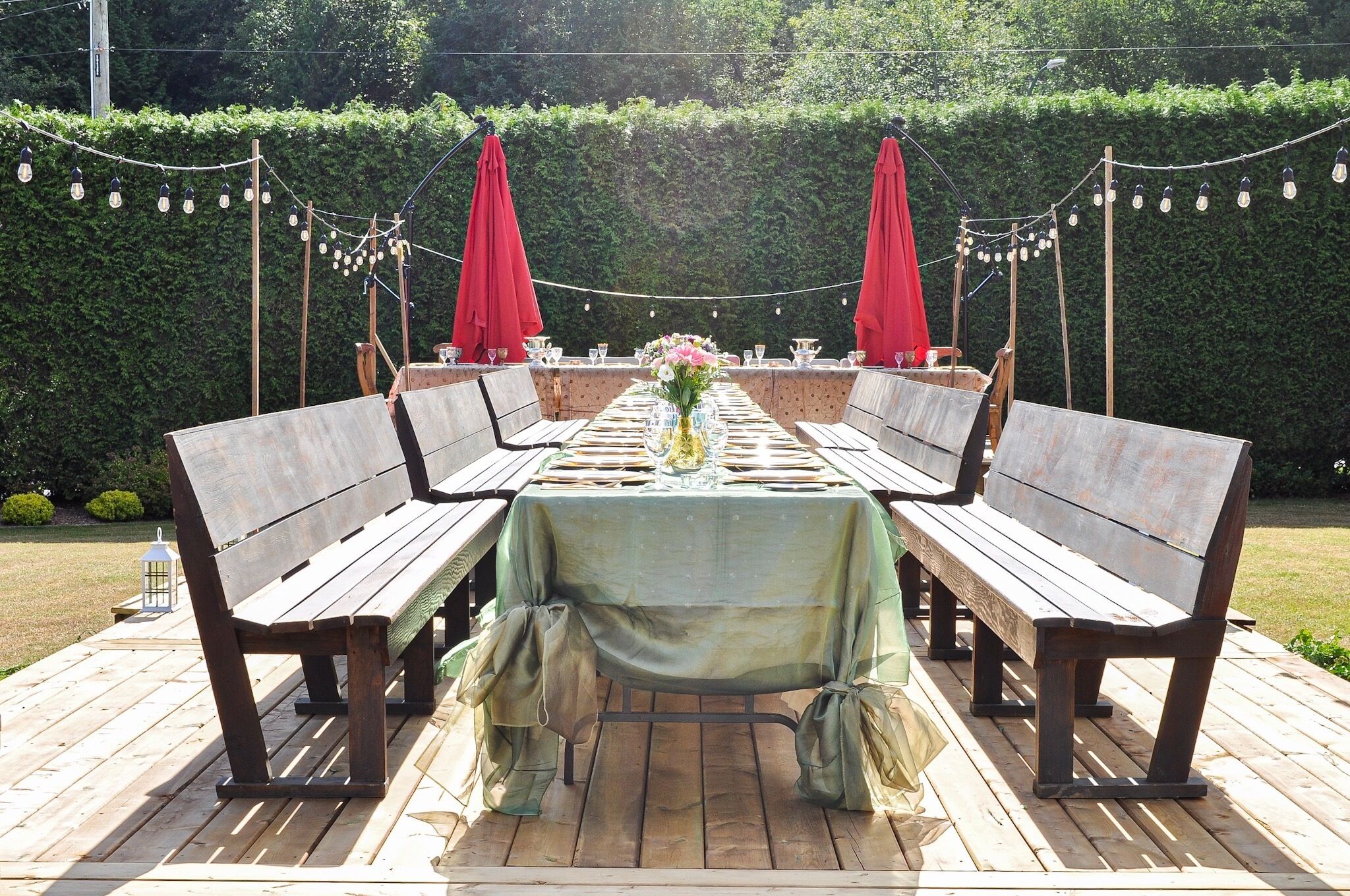 A wooden deck with a long table set for dining, surrounded by benches and red umbrellas.
