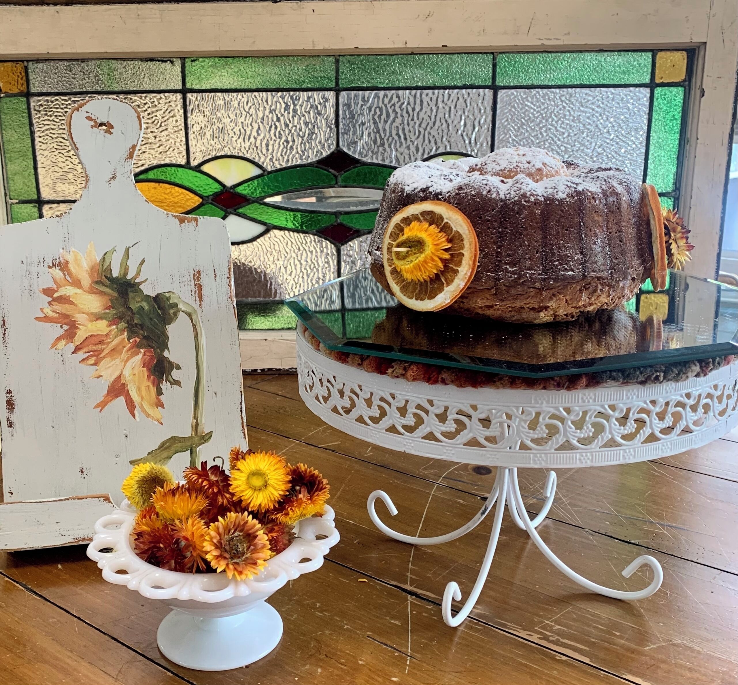 A cake on a glass stand with a floral arrangement beside it.
