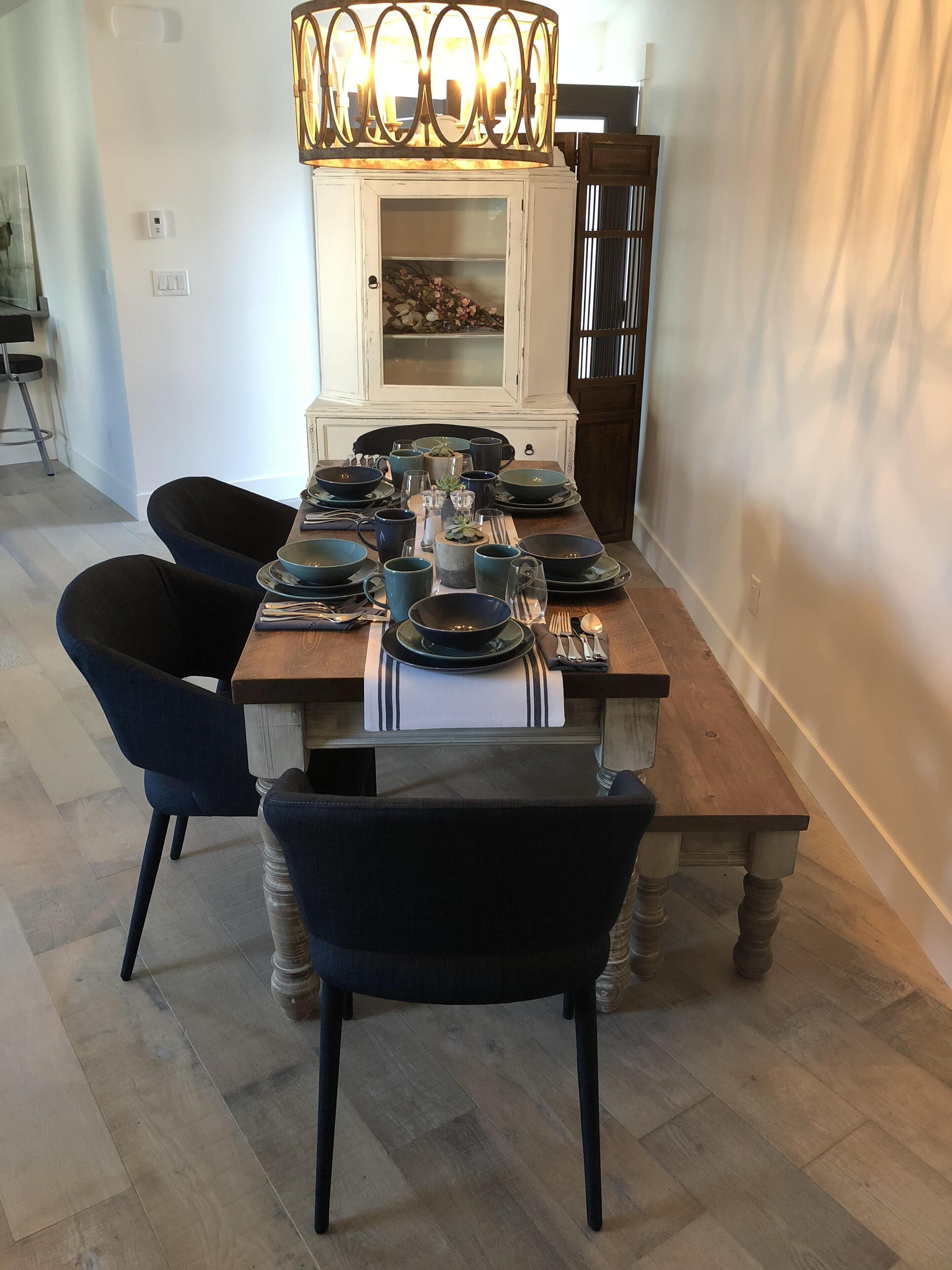 A dining area with a wooden table set for four, surrounded by dark upholstered chairs. A decorative light fixture hangs above the table, and a cabinet with glass doors is visible in the background.