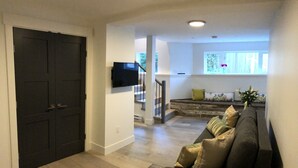 Uma sala de estar com um sofá de cor escura, uma TV de tela plana na parede e uma escada que leva a um andar superior.