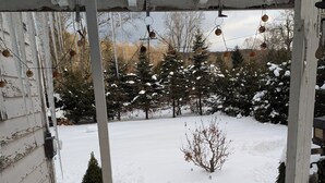 Patio nevado con una terraza de madera y vistas a árboles y una casa.