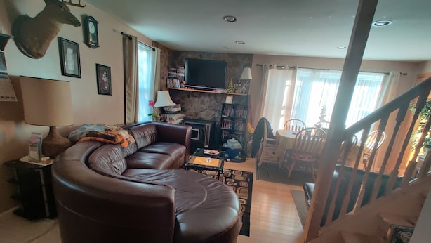 Una sala de estar con un sofá de cuero marrón en forma de L, un televisor de pantalla plana montado en una pared de piedra, una zona de comedor con sillas de madera y una mesa, y una escalera que conduce a otro nivel.