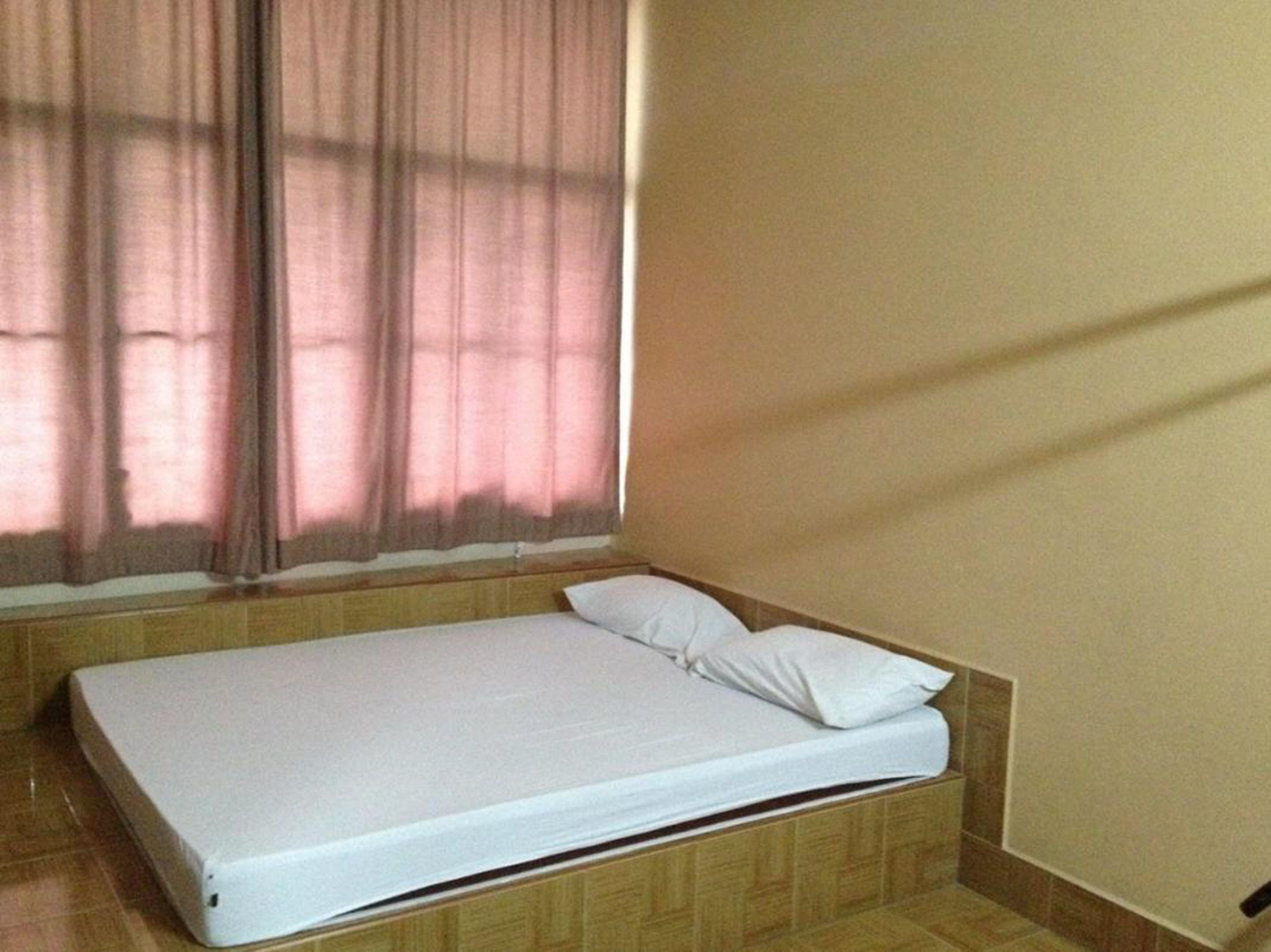 A single bed with a white mattress and pillow in a room with wooden paneling and a window with curtains.