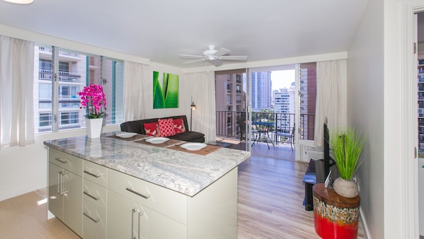 A modern kitchen with a marble island, a dining area with a sofa, and a balcony with a view of the city. - Designer Renovated, One Bedroom, Free Parking, Washer/dryer, Walk To Beach (Honolulu)