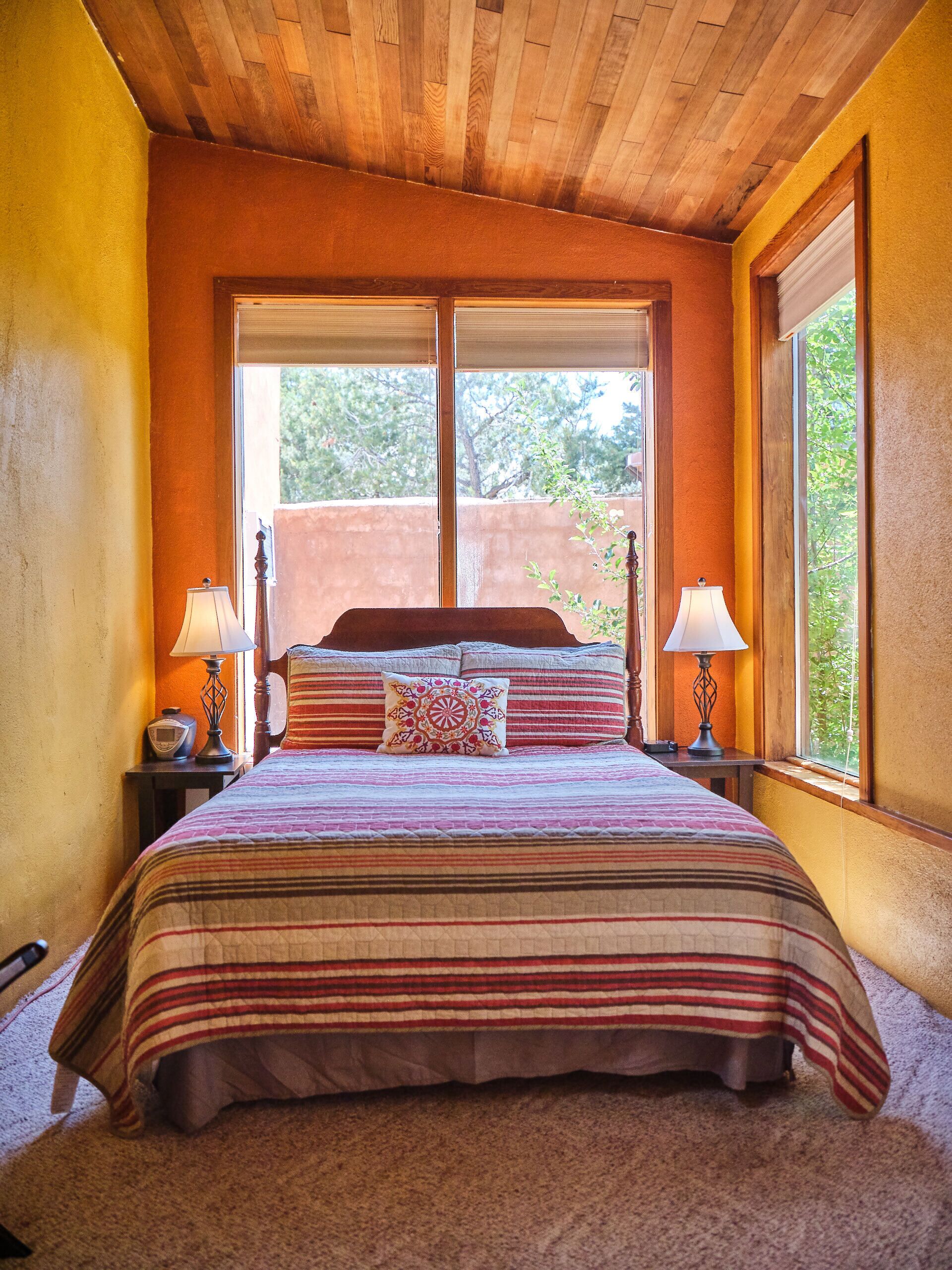 Un dormitorio con techo de madera, dos ventanas con persianas, una cama con ropa de cama a rayas y dos lámparas de noche.