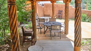A covered outdoor patio with a round table and chairs, surrounded by wooden columns. - Palace Of Miracles: Hot Tub, Lounge Spaces, +Casita, mtn views,5 courtyrds,6 FPs (El Prado)