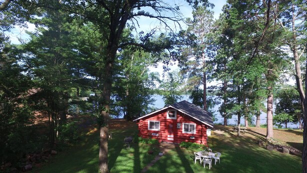 A red cabin surrounded by trees, with a grassy area and a picnic table nearby. - Peaceful Vintage 1920's Lakefront Red Log Cabin (Fifty Lakes)