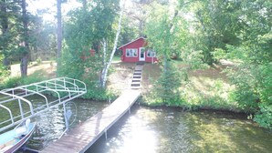 A wooden dock leading to a small red cabin surrounded by lush greenery and a calm body of water. - Peaceful Vintage 1920's Lakefront Red Log Cabin (Fifty Lakes)
